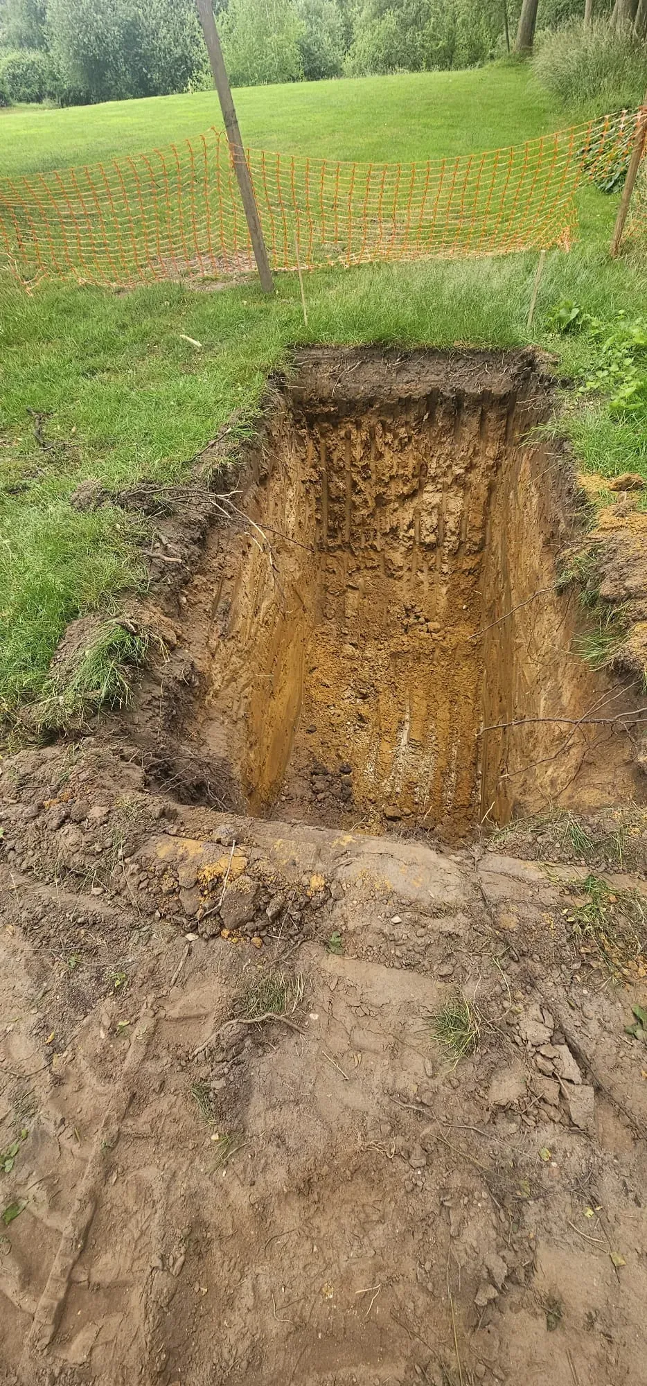 Deep excavation pit in a grassy area with orange safety fencing for groundworks or drainage installation.