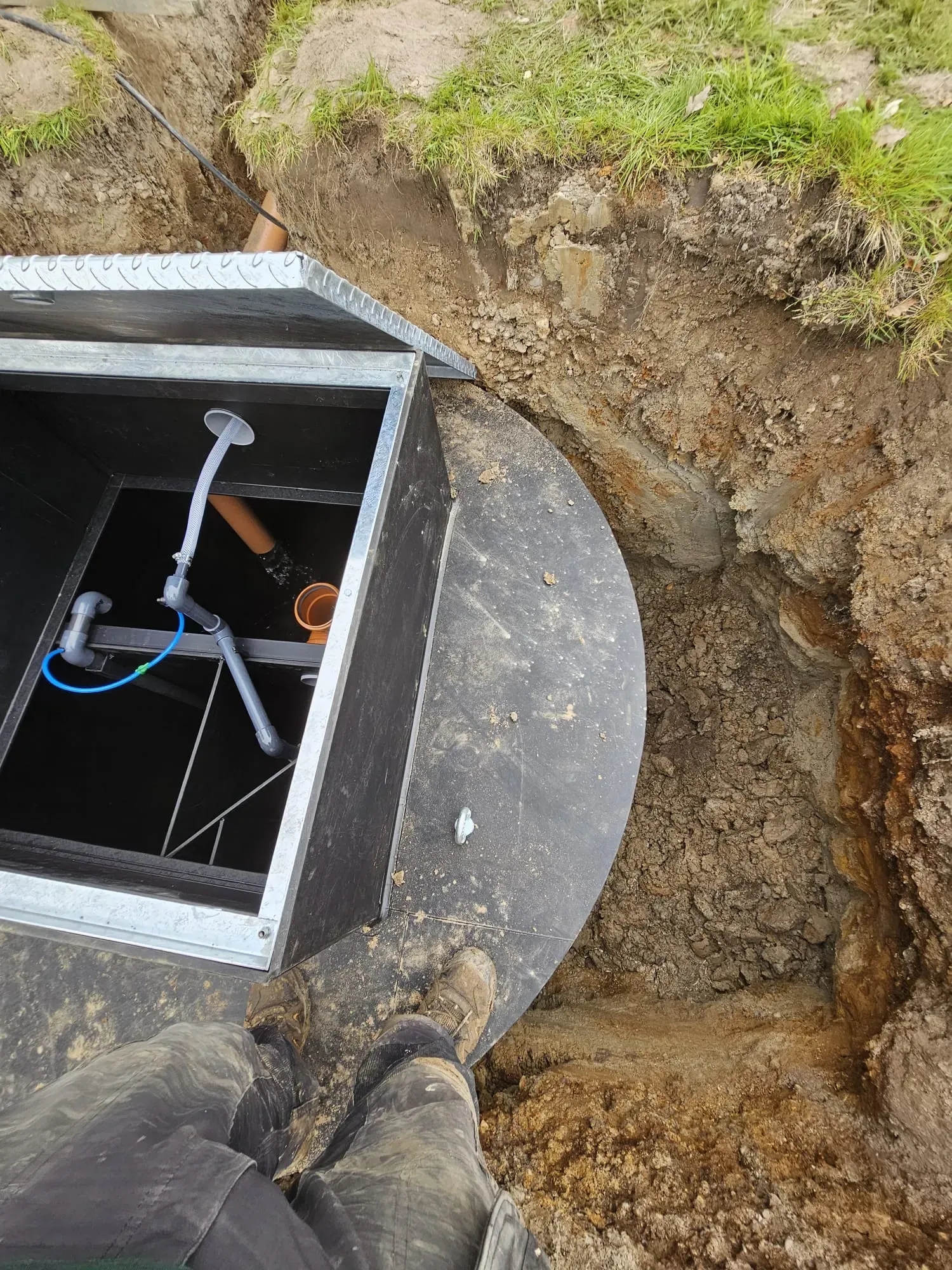 An engineer's muddy boots beside an open access chamber revealing internal pipes, during a septic tank or sewage treatment plant installation or repair in excavated earth.