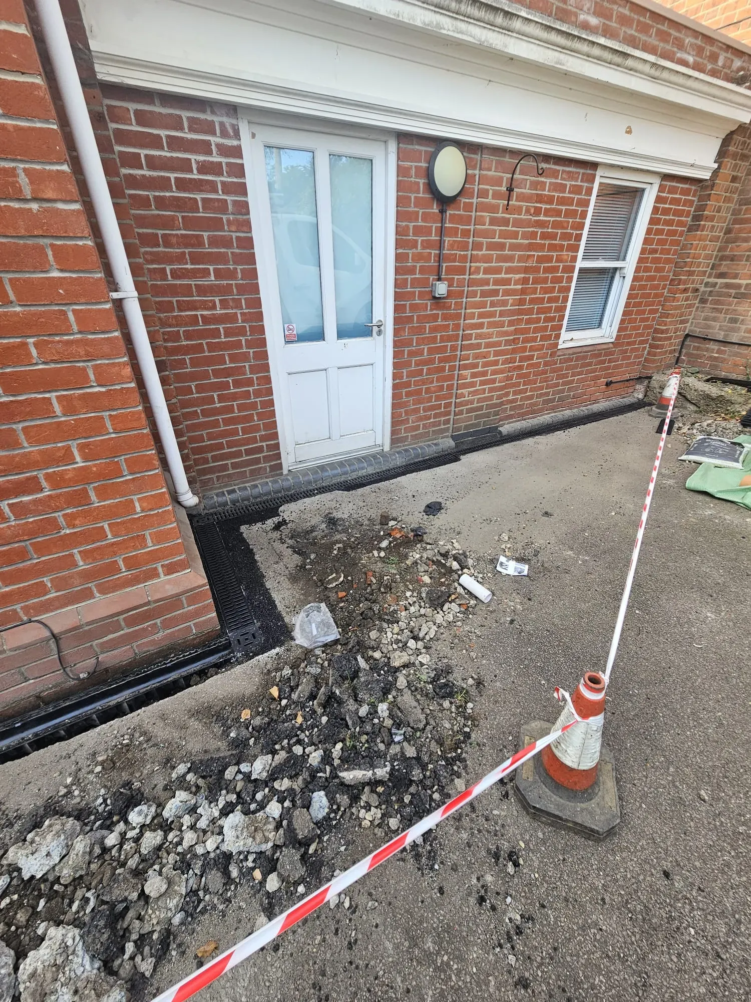 Image showing the installation of new channel drainage and groundwork activity next to a red brick building, with construction debris and a safety barrier.