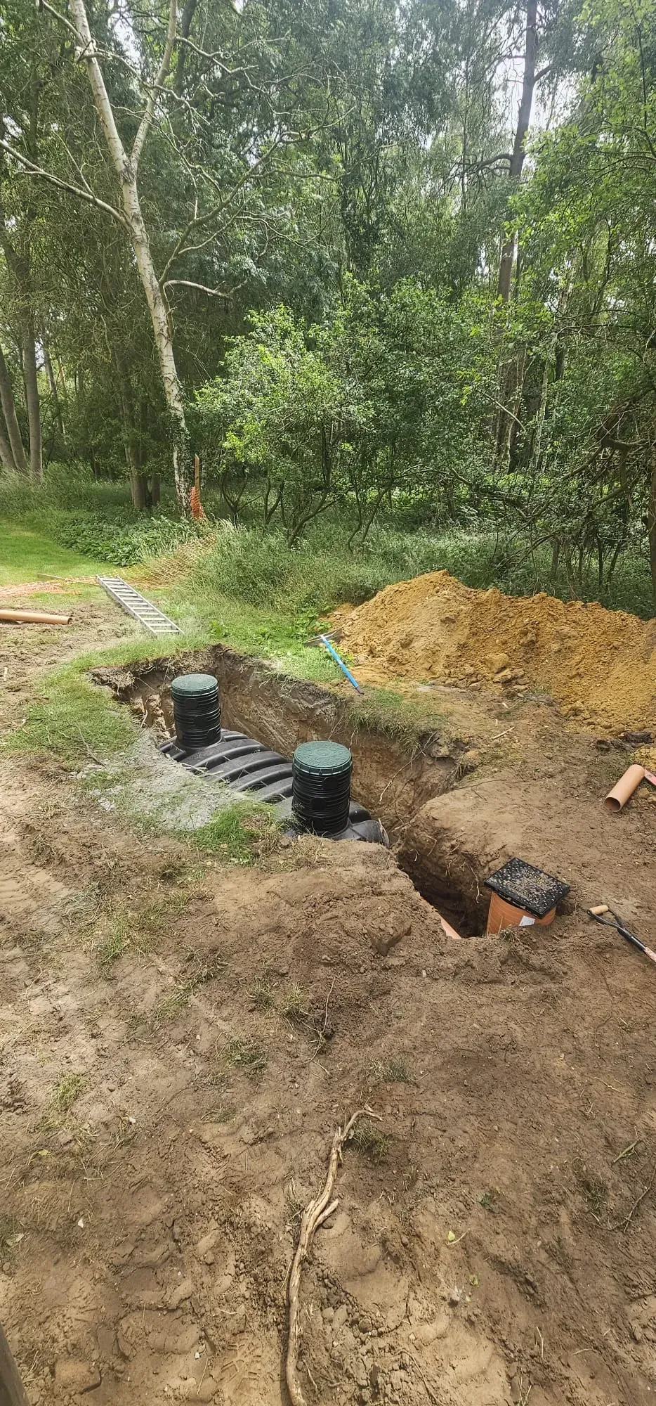 Septic tank or sewage treatment plant installation in progress in an open trench surrounded by dirt and trees.
