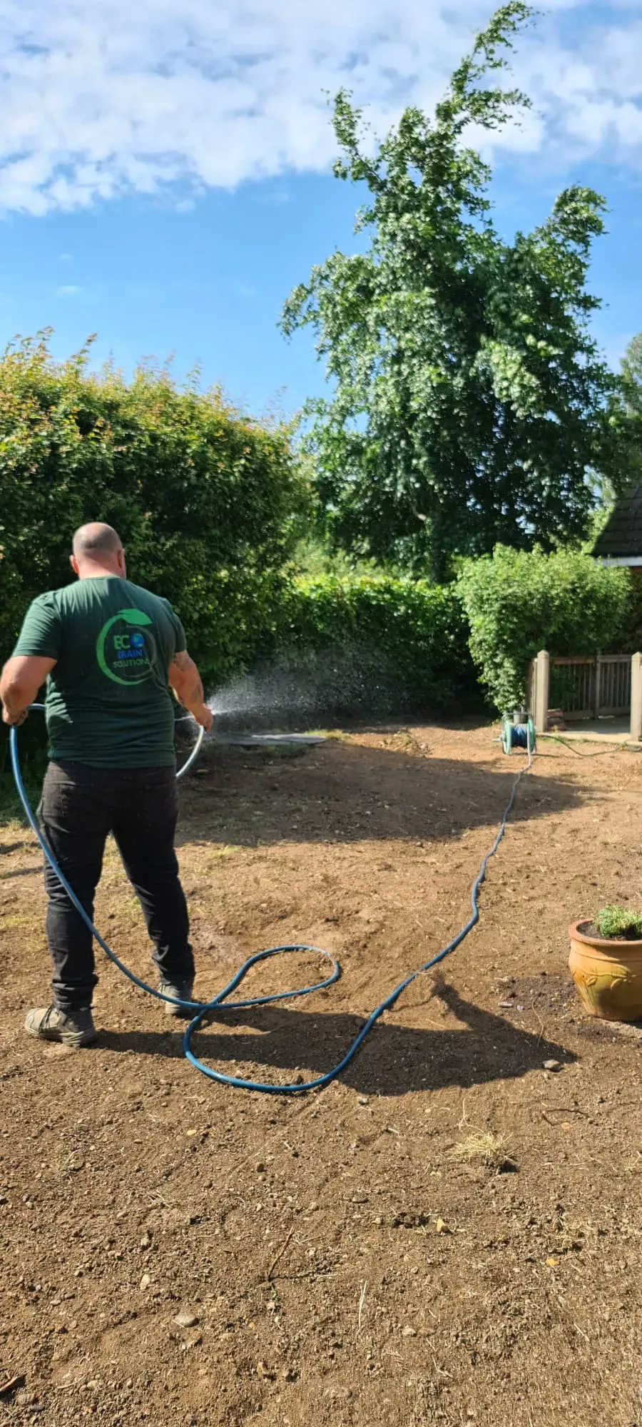 An Eco Drain Solutions Ltd technician in a green branded shirt watering a newly prepared soil area, likely for a groundworks or drainage installation project, with green trees and bushes in the background.