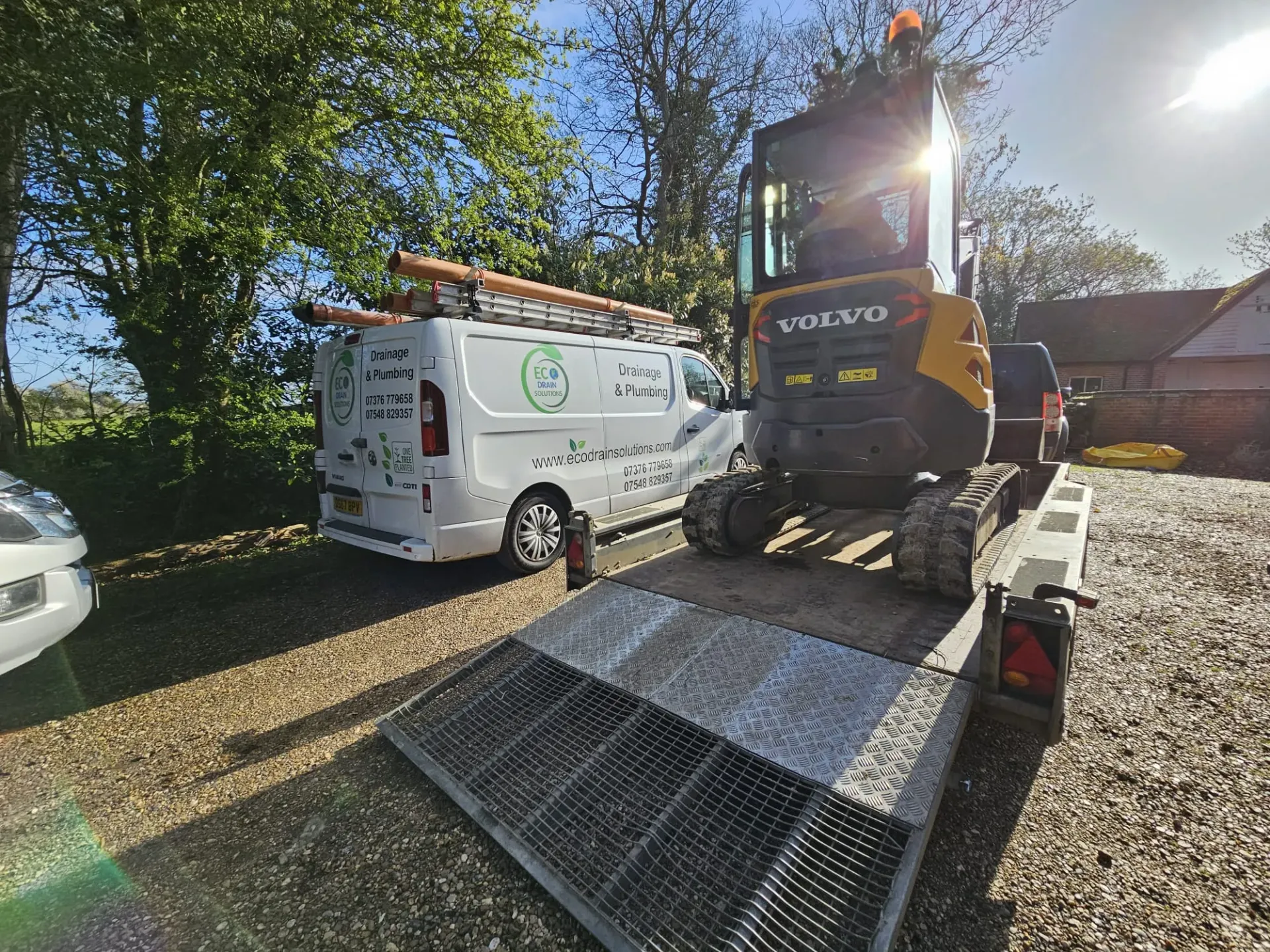 A white Eco Drain Solutions branded service van with roof-mounted pipes, parked next to a yellow Volvo mini excavator loaded on a trailer, on a gravel driveway under a bright sky.
